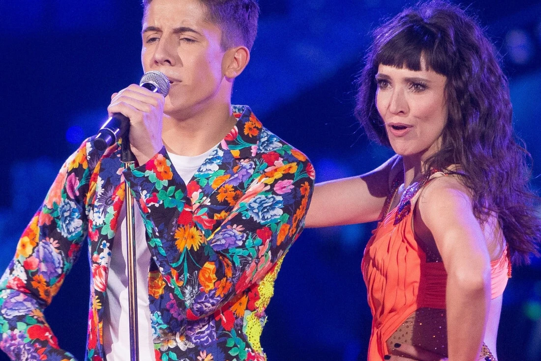 Dawid Kwiatkowski tańczył z Janją Leser w "Tańcu z gwiazdami" Dwójka młodych artystów występuje na scenie podczas koncertu. Mężczyzna w marynarce w kwiatowe wzory śpiewa do mikrofonu, obok niego kobieta w czerwonej sukni z szerokim uśmiechem obejmuje go ramieniem. Tło rozświetlają sceniczne światła w odcieniach n...