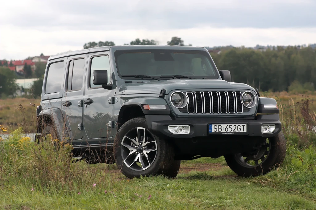 Jeep Wrangler Sahara to mieszanka starej, prawdziwej motoryzacji z nowoczesnymi rozwiązaniami Szary samochód terenowy Jeep Wrangler zaparkowany na polnej drodze, w tle drzewa i rozmyte budynki, auto wyeksponowane na pierwszym planie.