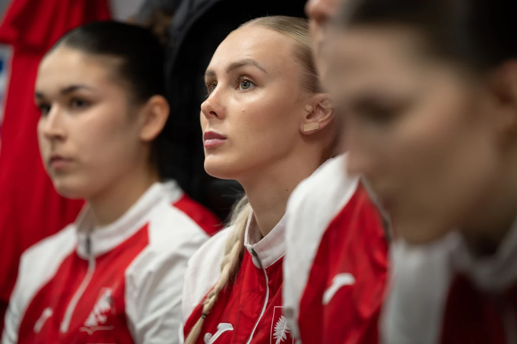 Grupa młodych kobiet w sportowych czerwono-białych strojach z logo orła, skupione, zwrócone ku jednemu punktowi, w tle widoczne inne postaci niewyraźnie.