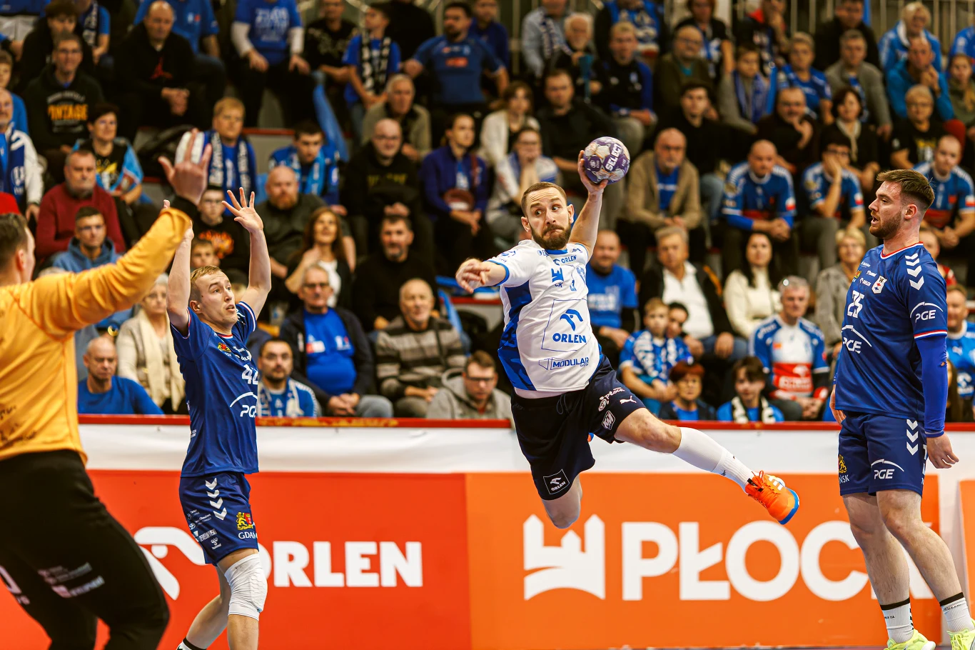 Zawodnik drużyny piłki ręcznej w białej koszulce wyskakuje w powietrze, celując piłką w stronę bramkarza drużyny przeciwnej ubranego na żółto, wokół niego kilku graczy w niebieskich strojach, a w tle widoczna publiczność na trybunach.