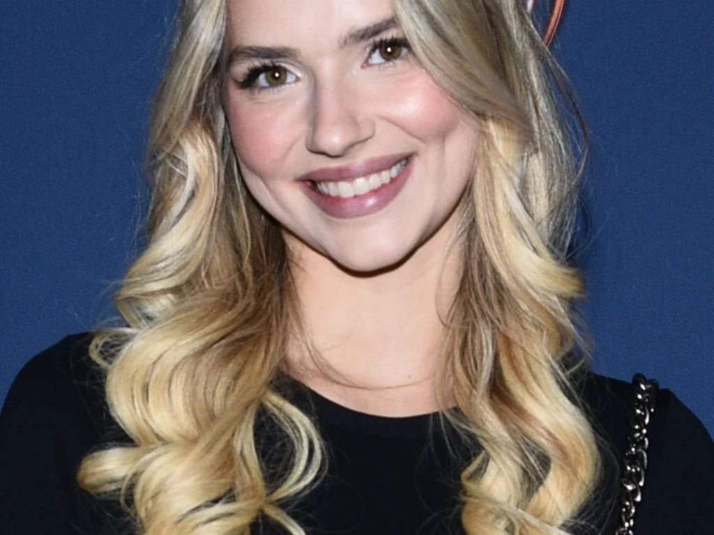 Młoda kobieta o długich, falowanych blond włosach i jasnej karnacji uśmiecha się, patrząc wprost w obiektyw. Ubrana w czarną bluzkę, na ramieniu ma torebkę na łańcuszku. Za nią jednolite, ciemnoniebieskie tło.