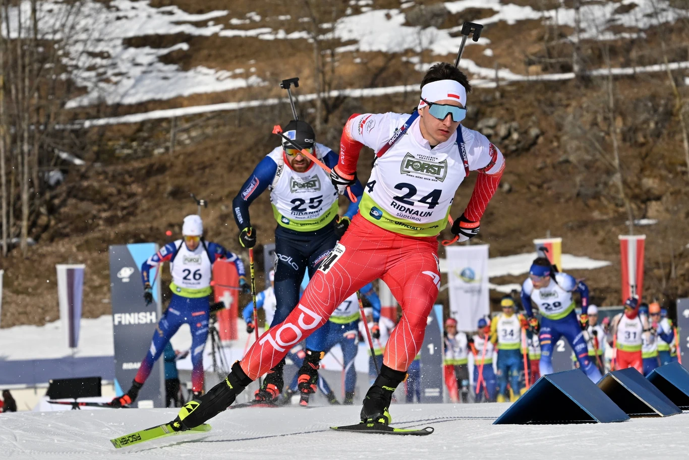 Wojciech Skorusa Zawodnicy biorący udział w wyścigu biathlonowym na śniegu, ubrani w sportowe stroje i numery startowe, z karabinami na plecach, na tle zimowego krajobrazu i licznych flag.