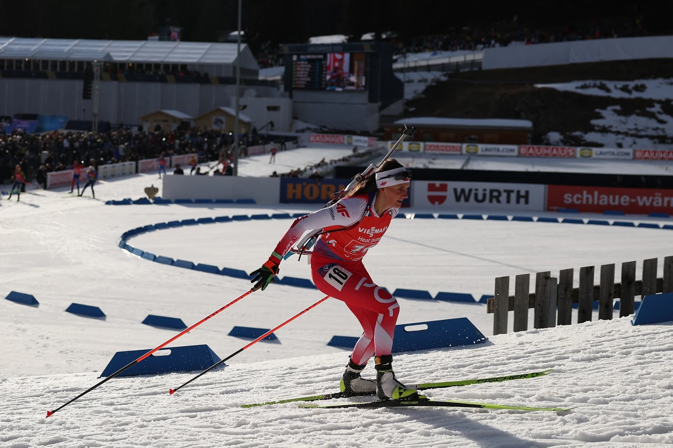 Joanna Jakieła Zawodniczka biathlonowa w czerwono-białym stroju podczas dynamicznej jazdy na nartach, z widocznym karabinem na plecach; w tle trybuny pełne widzów oraz elementy infrastruktury sportowej i reklamowej.