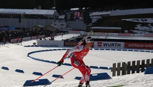 Zawodniczka biathlonowa w czerwono-białym stroju podczas dynamicznej jazdy na nartach, z widocznym karabinem na plecach; w tle trybuny pełne widzów oraz elementy infrastruktury sportowej i reklamowej.