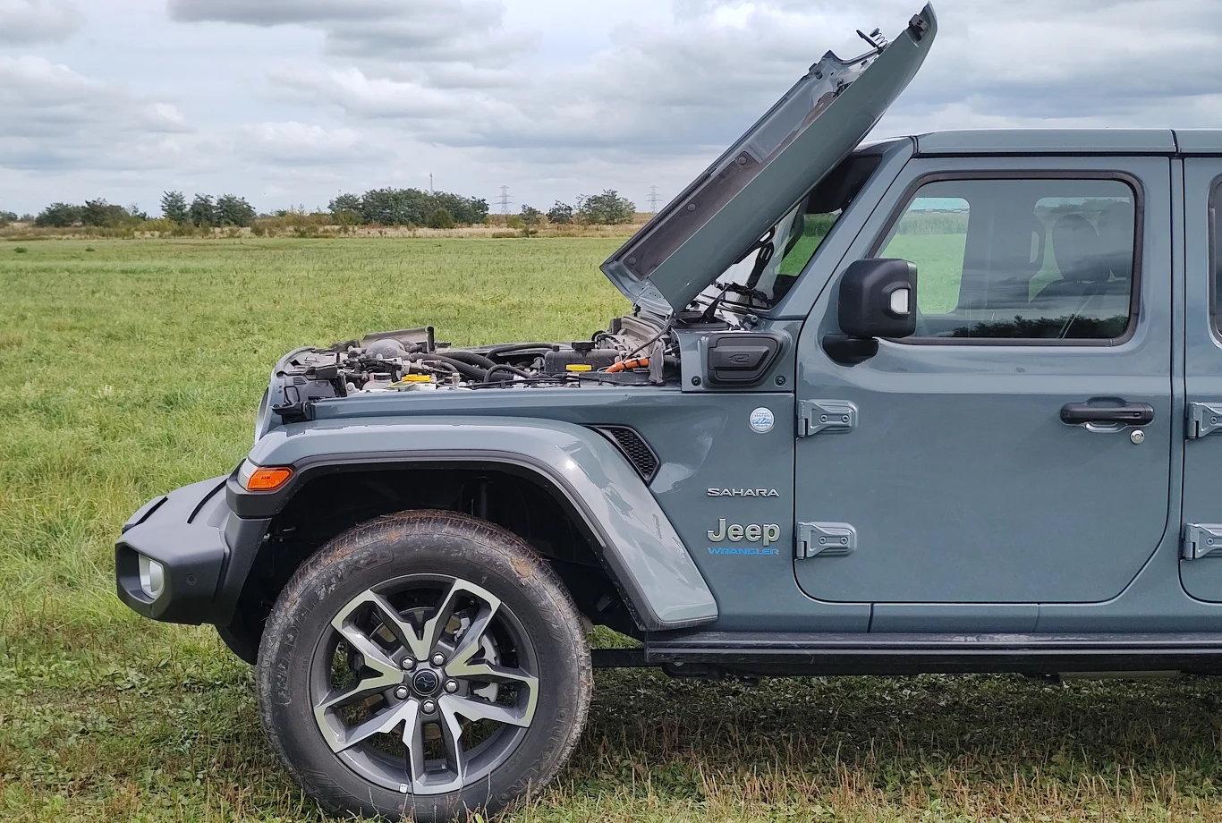 Klapę silnika można oprzeć o dach, co zapewnia doskonały dostęp do silnika. Podczas manewrów trzeba uważać na mocno wystający zderzak Samochód terenowy Jeep Wrangler zaparkowany na łące z otwartą maską silnika, widoczna przednia część pojazdu oraz szczegóły mechaniczne pod maską. Zielone pole i pochmurne niebo w tle.