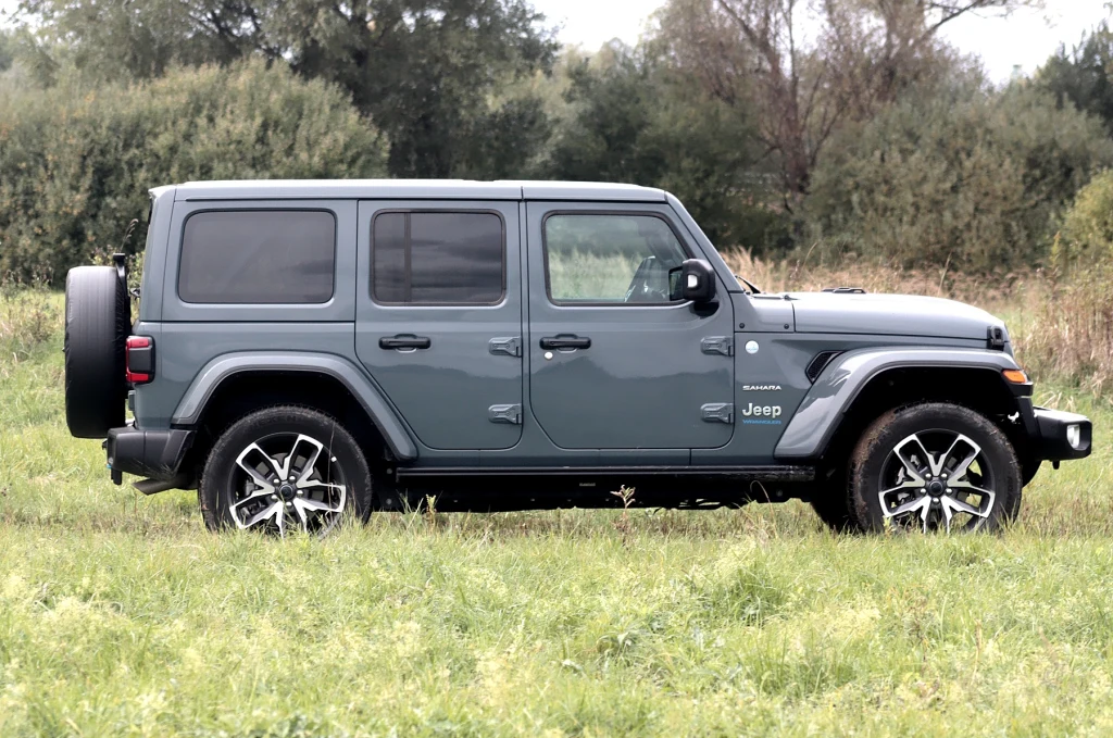 Szyba Wrangler jest niewielka i umieszczona pionowo Samochód terenowy Jeep Wrangler w zielonym kolorze ustawiony na trawie, w otoczeniu zieleni oraz drzew, widok z boku.