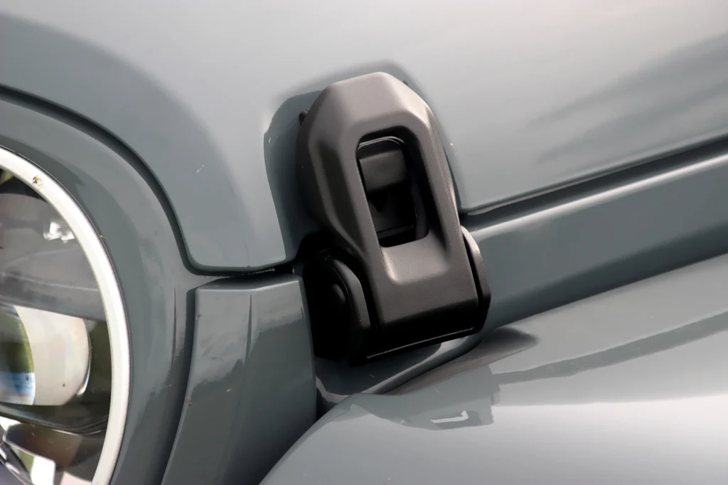 Blokady, które trzeba zwolnić, by dostać się do silnika Zbliżenie na maskę samochodu terenowego, na której widoczny jest masywny czarny zatrzask w okolicach reflektora oraz szara karoseria.