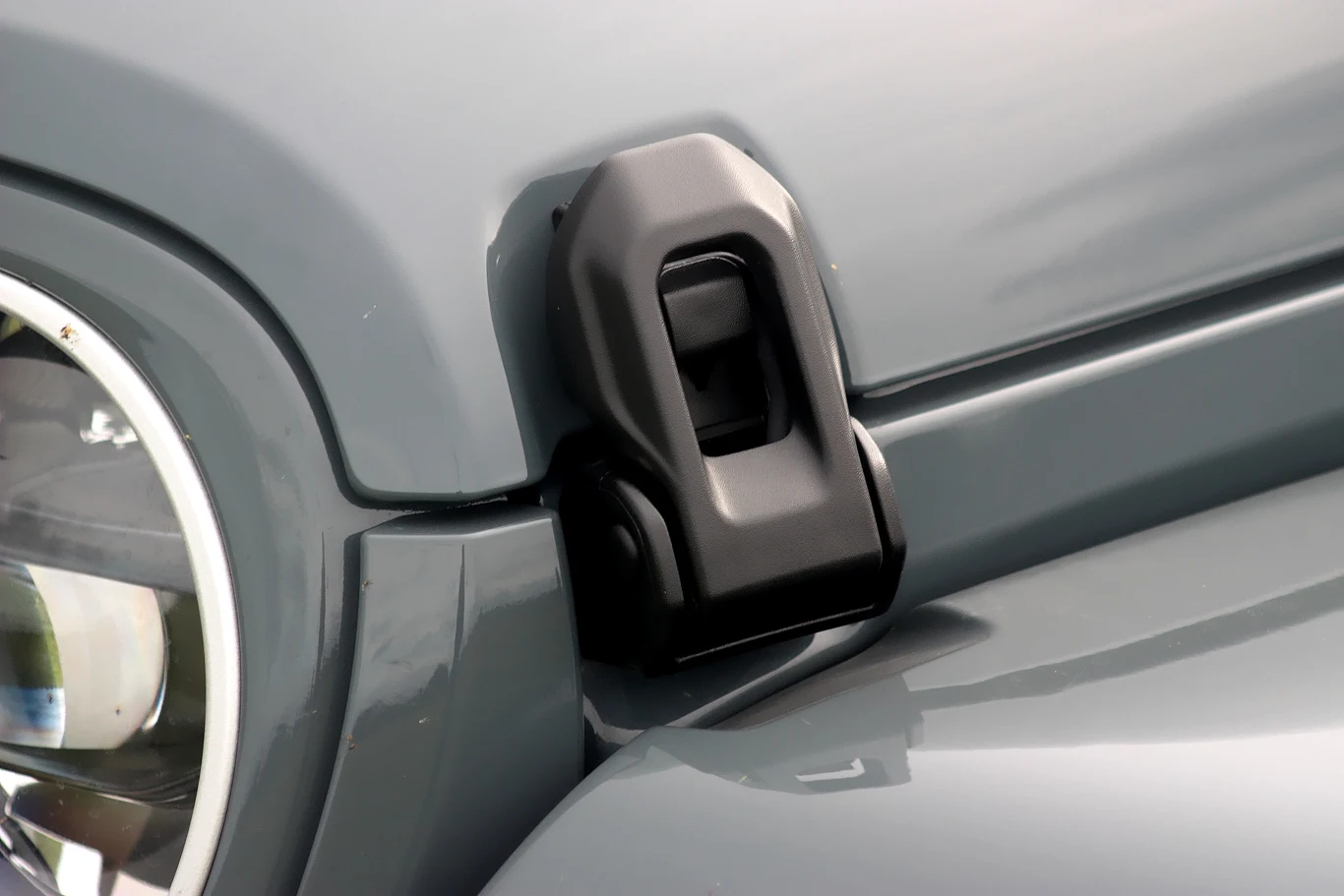 Blokady, które trzeba zwolnić, by dostać się do silnika Zbliżenie na maskę samochodu terenowego, na której widoczny jest masywny czarny zatrzask w okolicach reflektora oraz szara karoseria.