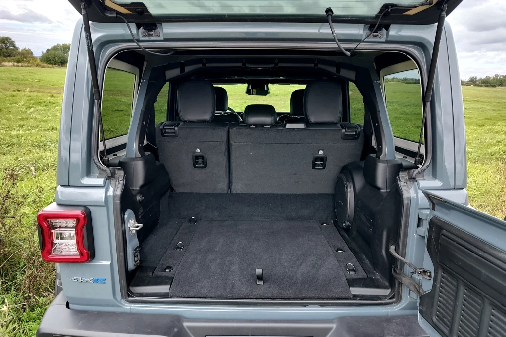 Bagażnik Wrangler ma spore rozmiary. Widoczny potężny pałąk przeciwkapotażowy, wokół którego obudowanego nadwozie Otwarty bagażnik samochodu terenowego z widocznym przestronnym wnętrzem oraz złożonymi tylnymi siedzeniami, znajdujący się na tle zielonego pola i nieba z chmurami.