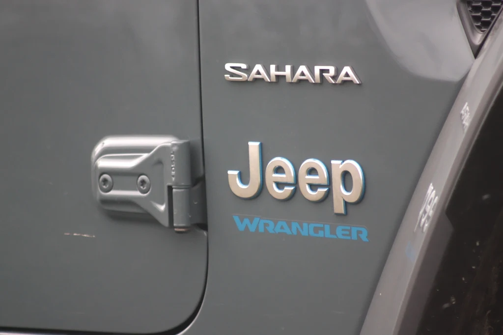 Drzwi można łatwo odkręcić dzięki zewnętrznym zawiasom Logo Jeep oraz napisy Sahara i Wrangler na ciemnoszarej powierzchni samochodu terenowego, widoczna także część mechanizmu drzwi.