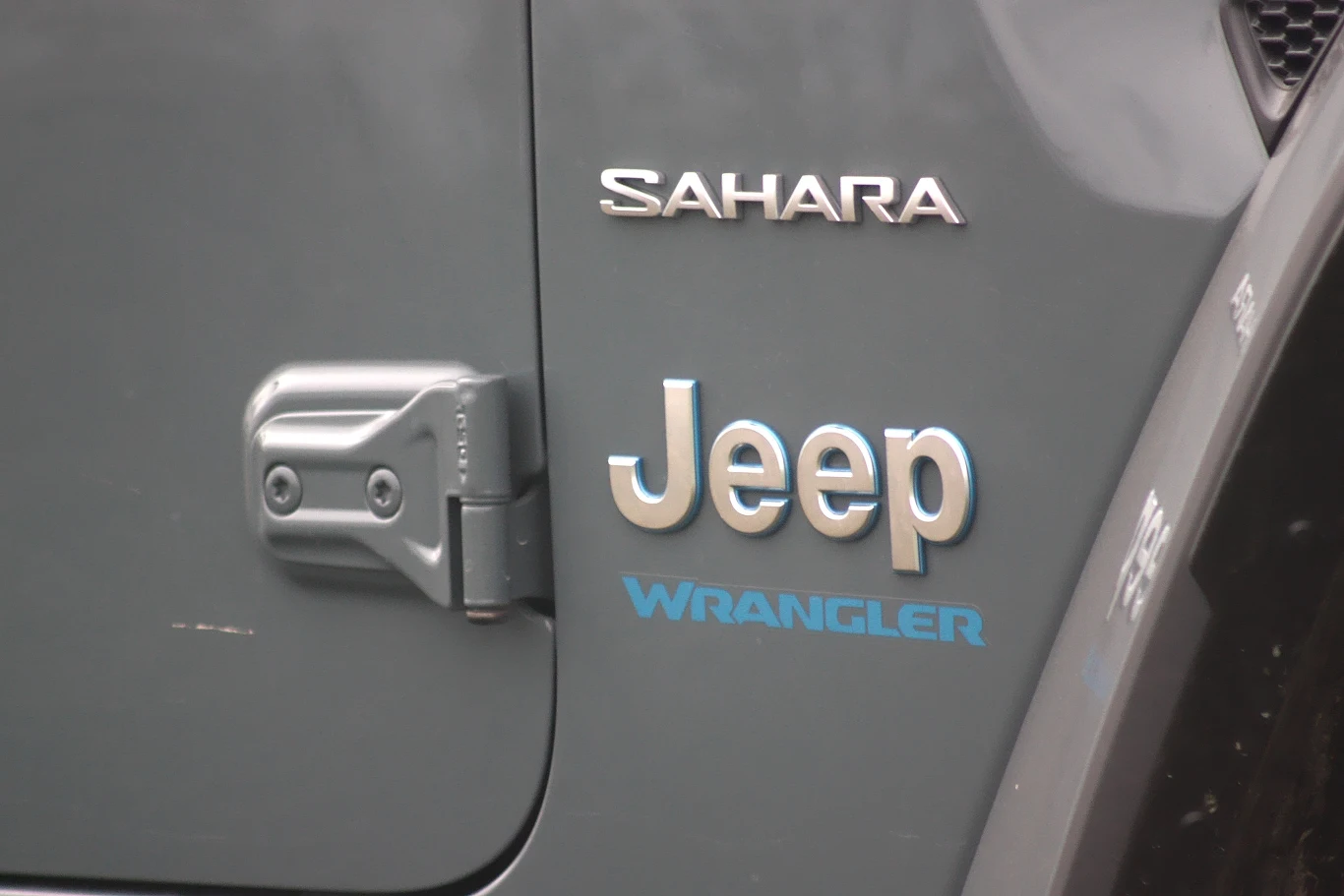 Drzwi można łatwo odkręcić dzięki zewnętrznym zawiasom Logo Jeep oraz napisy Sahara i Wrangler na ciemnoszarej powierzchni samochodu terenowego, widoczna także część mechanizmu drzwi.