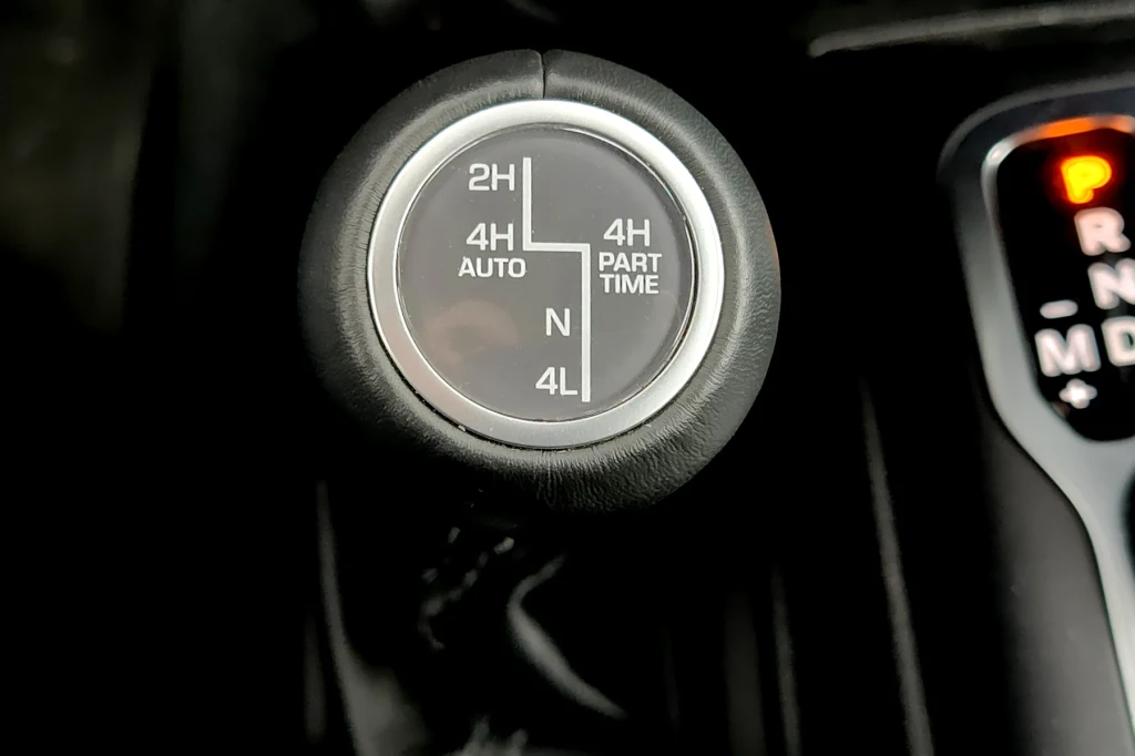 Dźwignia sterowania napędem Jeepa Wranglera Gałka zmiany trybu napędu w samochodzie terenowym z oznaczeniami różnych trybów jazdy, widoczna również część dźwigni automatycznej skrzyni biegów ze świecącą literą P.