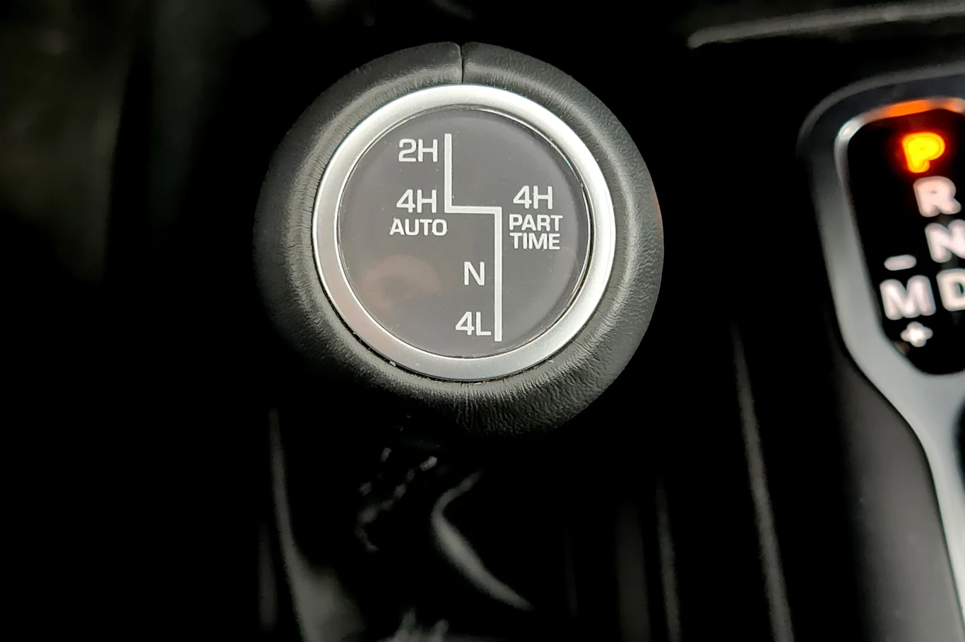 Dźwignia sterowania napędem Jeepa Wranglera Gałka zmiany trybu napędu w samochodzie terenowym z oznaczeniami różnych trybów jazdy, widoczna również część dźwigni automatycznej skrzyni biegów ze świecącą literą P.