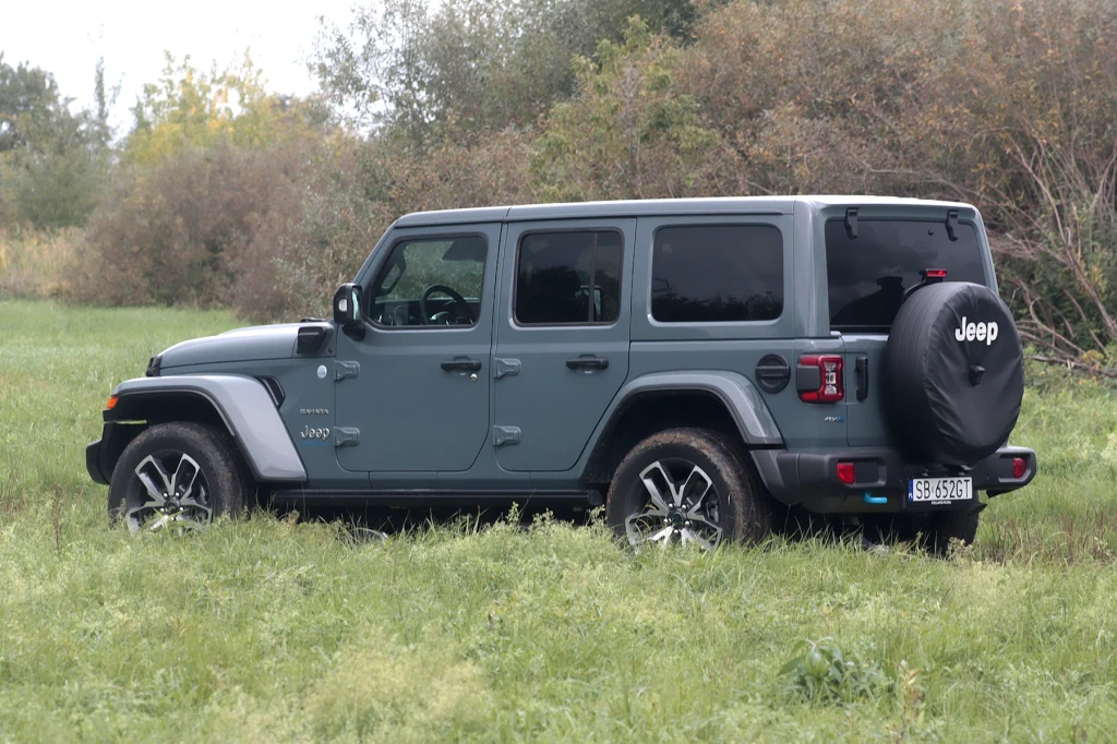 Szkoda, że normy emisji spalin pokonały Jeepa Wranglera Szary samochód terenowy marki Jeep stoi na trawie, otoczony zielenią i drzewami, w naturalnym środowisku, prezentując się w ujęciu z boku i tyłu.