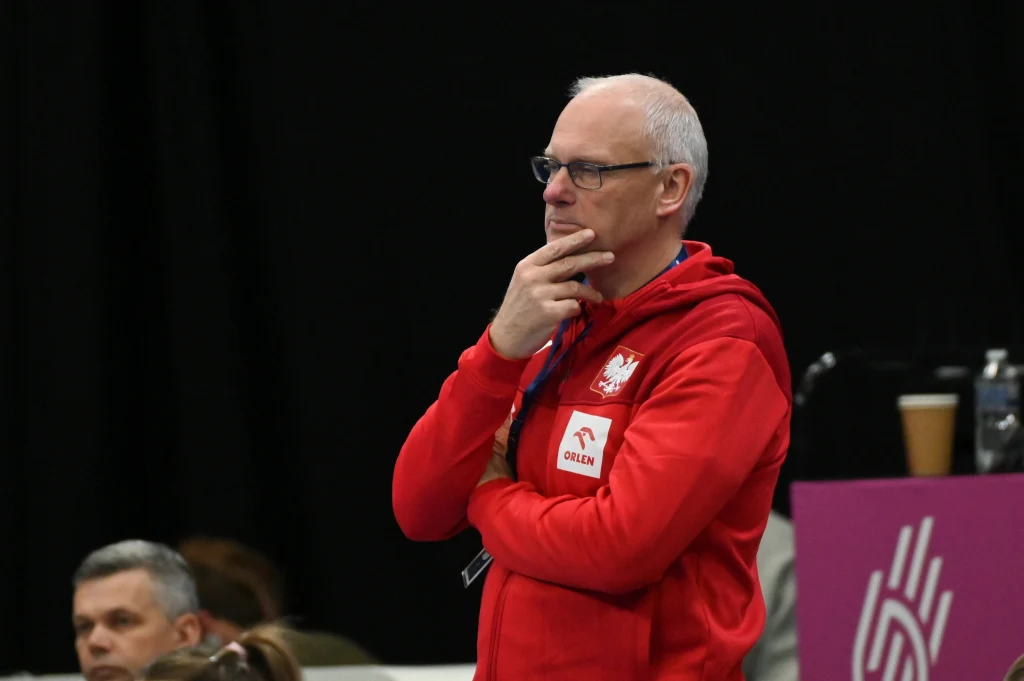 Trener w czerwonej bluzie z polskim godłem i logo sponsora zamyślony podczas sportowego wydarzenia, w tle widoczni inni uczestnicy oraz kubek na stole.