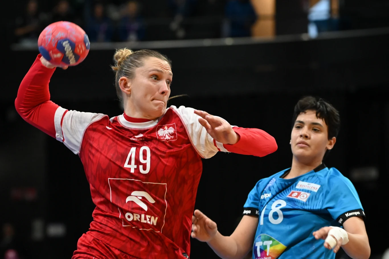 Karolina Kochaniak Zawodniczka w czerwonym stroju z numerem 49 wykonuje dynamiczny rzut piłką ręczną, obok znajduje się przeciwniczka w niebieskim stroju z numerem 8, obie skoncentrowane na grze.