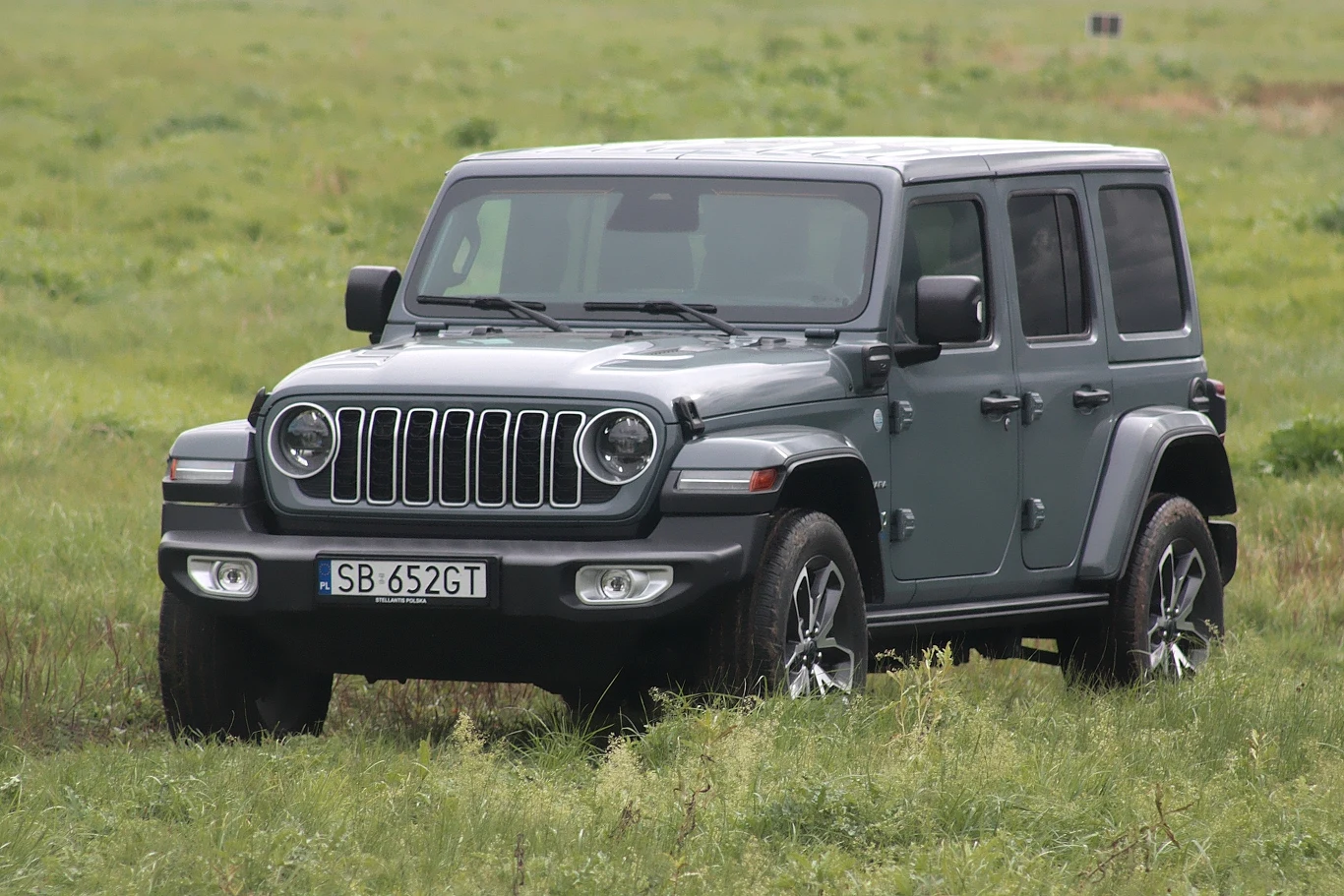 Jeep Wrangler Sahara Szary samochód terenowy Jeep Wrangler z polską tablicą rejestracyjną SB 652GT stoi na zielonej, trawiastej łące. W tle widoczny rozległy teren bez zabudowań.