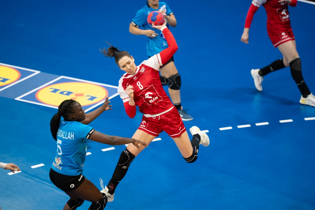 Zawodniczka w czerwonym stroju oddaje rzut podczas meczu piłki ręcznej, próbując zdobyć bramkę, podczas gdy obrończyni w niebieskiej koszulce stara się ją zablokować. Tło stanowią elementy boiska oraz inne zawodniczki.