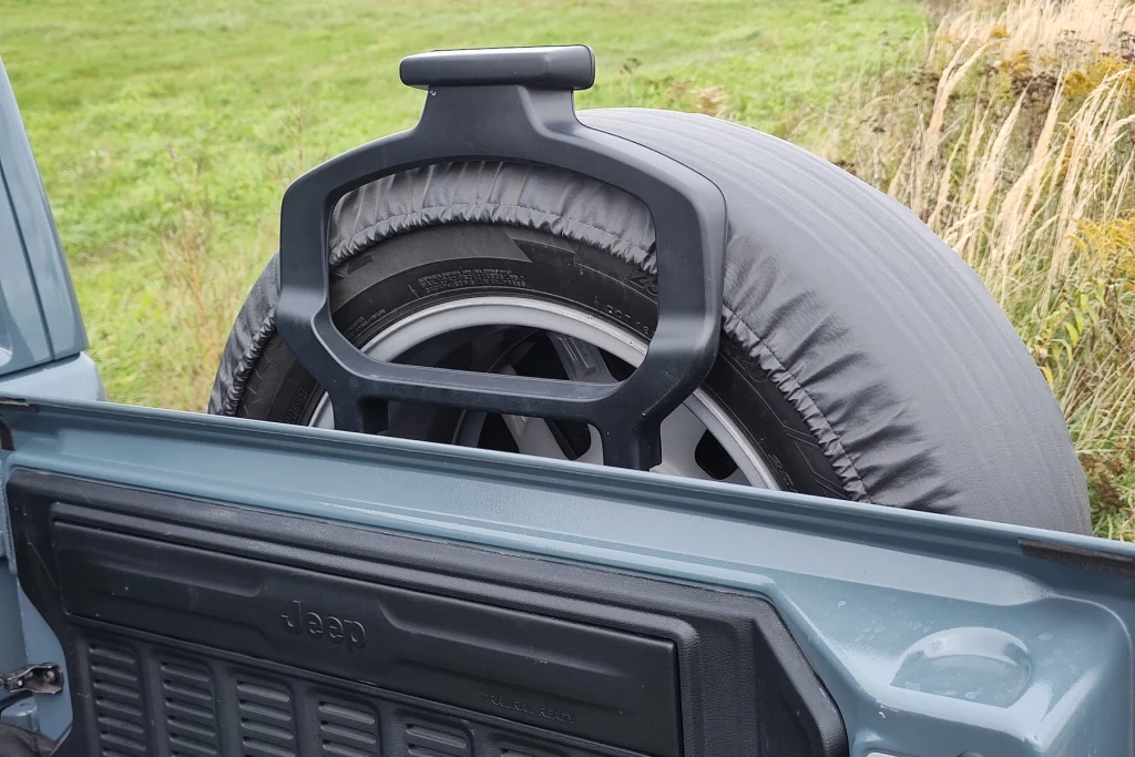 Jeep Wrangler Sahara Bagażnik samochodu terenowego z zamontowanym kołem zapasowym zabezpieczonym pokrowcem oraz elementem uchwytu na kole, wokół widoczna trawa i fragment pola.