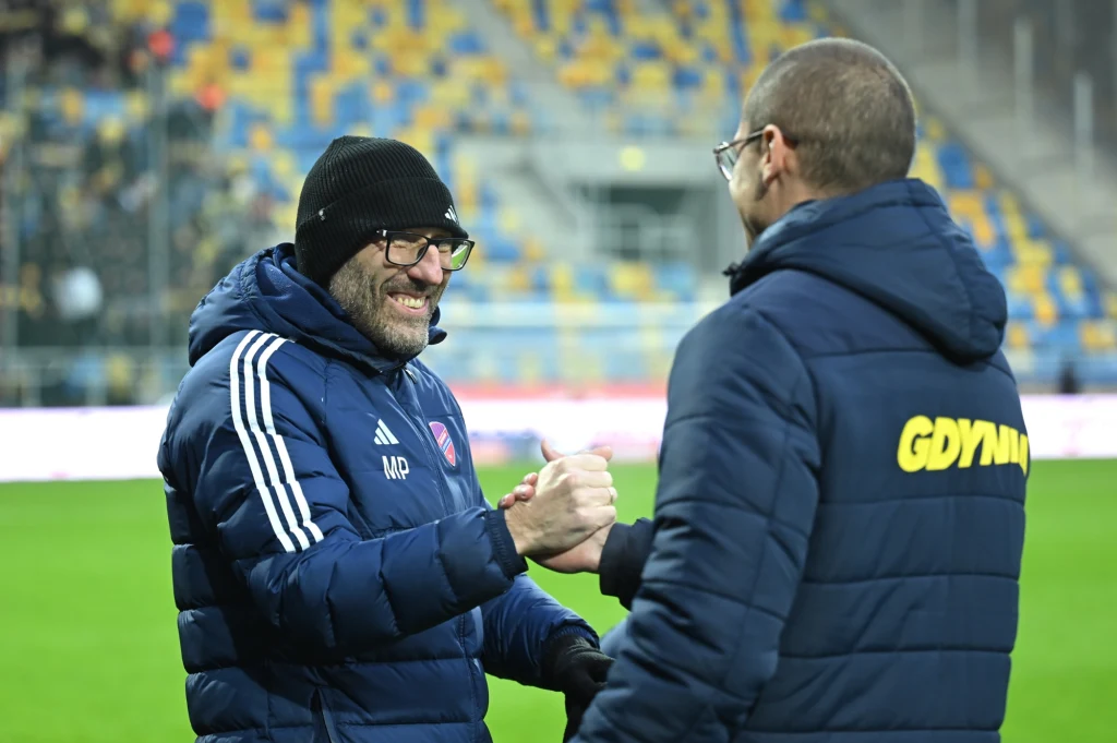 Marek Papszun (z lewej) wygląda, jakby już przed meczem wiedział, co czeka jego niedawnego asystenta Dawida Szwargę Dwóch mężczyzn ubranych w zimowe kurtki i czapki ściska sobie dłonie na tle stadionu piłkarskiego, jeden z nich ubrany jest w kurtkę z napisem 'GDYNIA', atmosfera jest przyjacielska i sportowa.