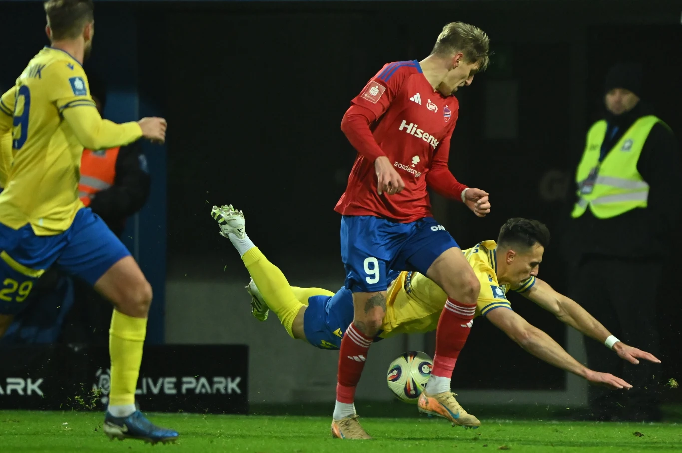 Dwóch piłkarzy walczy o piłkę podczas meczu piłki nożnej, jeden z nich upadł na murawę, natomiast sędzia techniczny i inny zawodnik pozostają w tle. Napięcie i rywalizacja sportowa skupione są w centrum akcji.