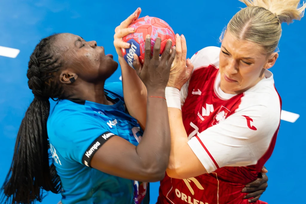 Moment walki o piłkę podczas meczu piłki ręcznej kobiet, dwie zawodniczki z różnych drużyn ścierają się przy próbie przejęcia piłki; dynamiczna i intensywna ekspresja na twarzach zawodniczek ukazuje zaciętą rywalizację sportową.