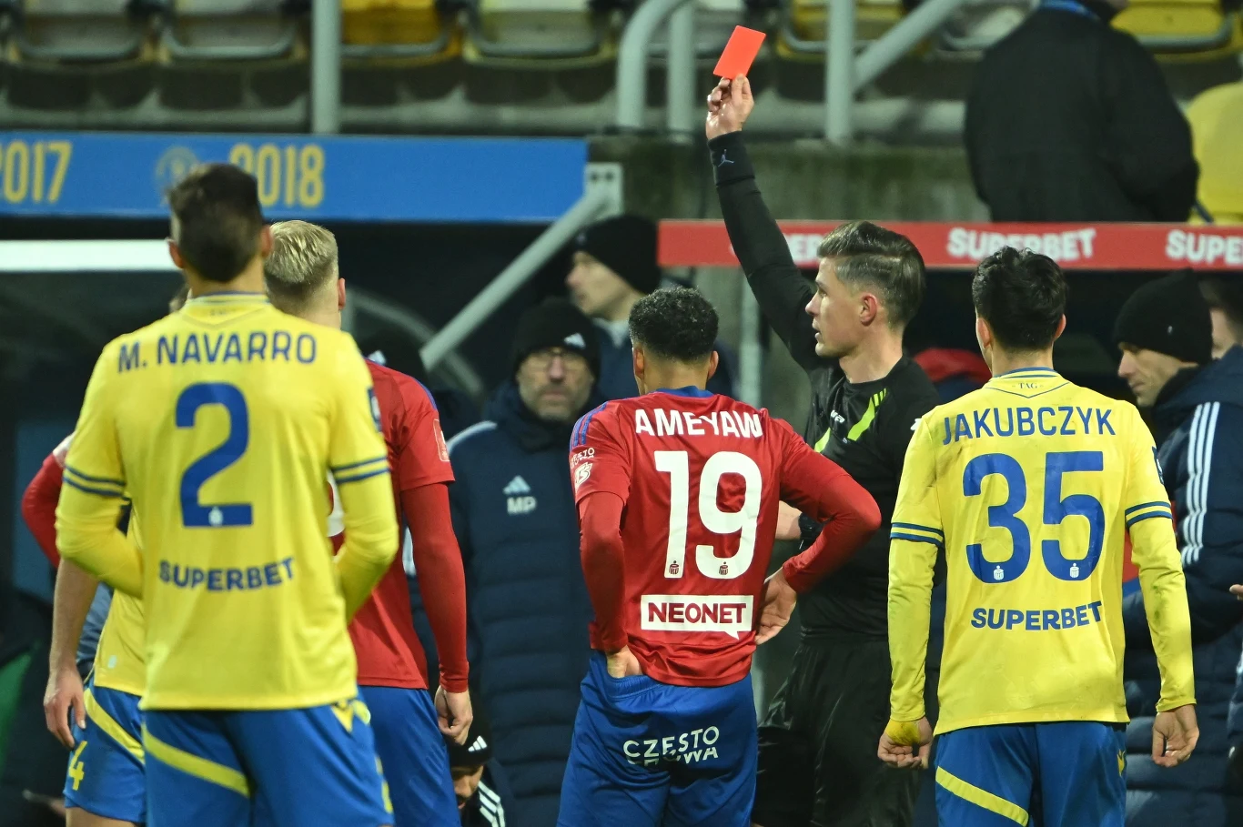 Sędzia pokazuje czerwoną kartkę jednemu z zawodników podczas meczu piłki nożnej, wokół niego kilku piłkarzy z drużyn w żółtych i czerwonych strojach, całość rozgrywa się na stadionie.