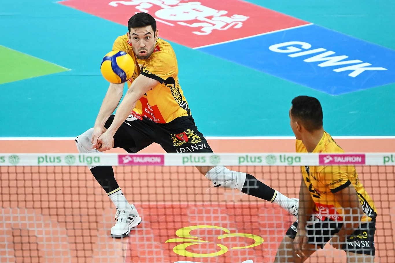 Siatkarz w żółto-czarnym stroju gdańskiej drużyny odbija piłkę techniką dolną podczas meczu siatkówki na hali. W tle widoczny drugi zawodnik w tej samej drużynie oraz fragmenty boiska z wyraźnie oznaczonymi liniami i logotypami sponsorów.