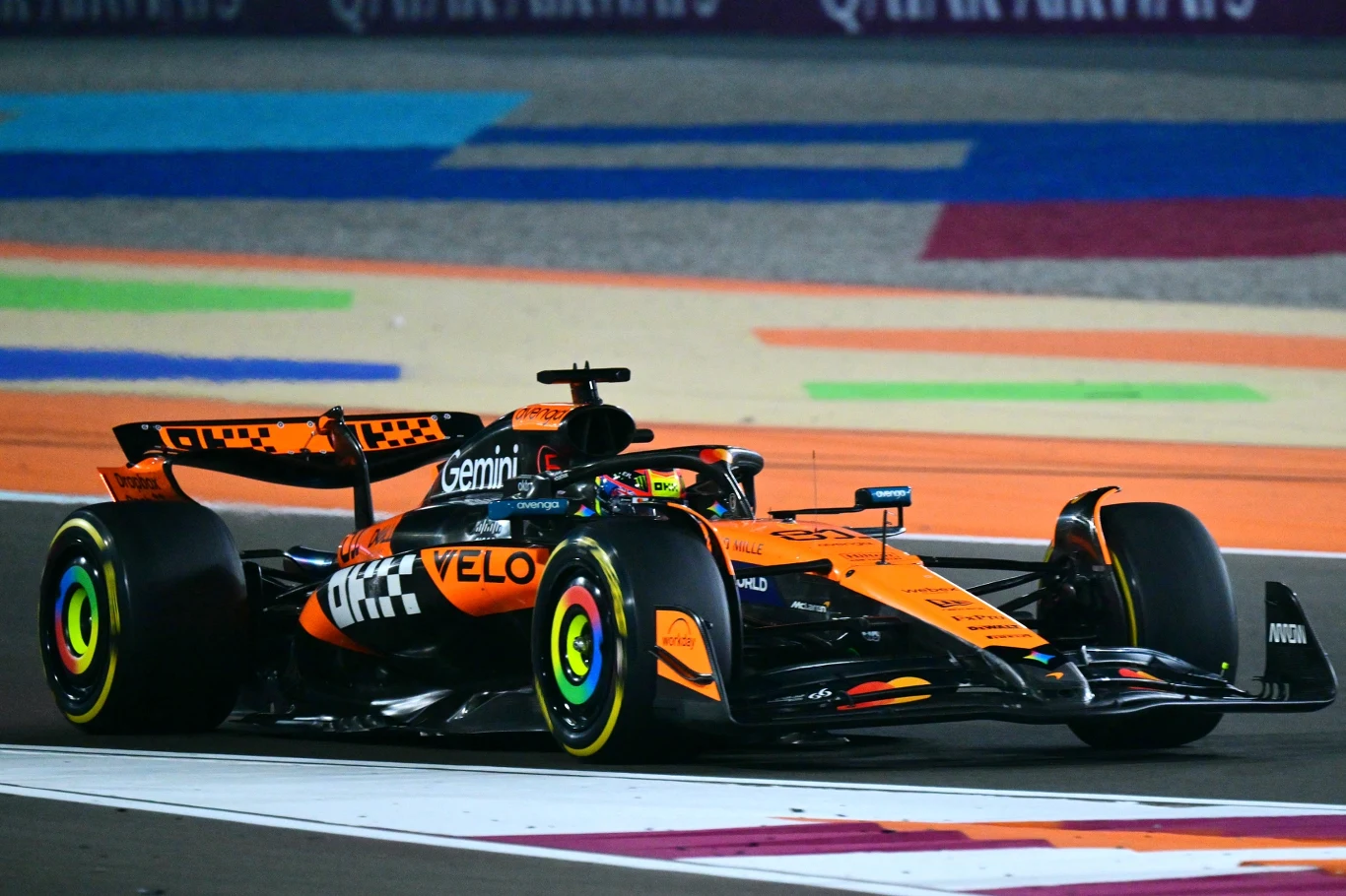 Oscar Piastri Formułowy bolid wyścigowy McLarena o pomarańczowo-czarnym malowaniu w trakcie dynamicznej jazdy na torze, otoczony kolorowymi liniami toru oraz elementami reklamy na tle asfaltu.