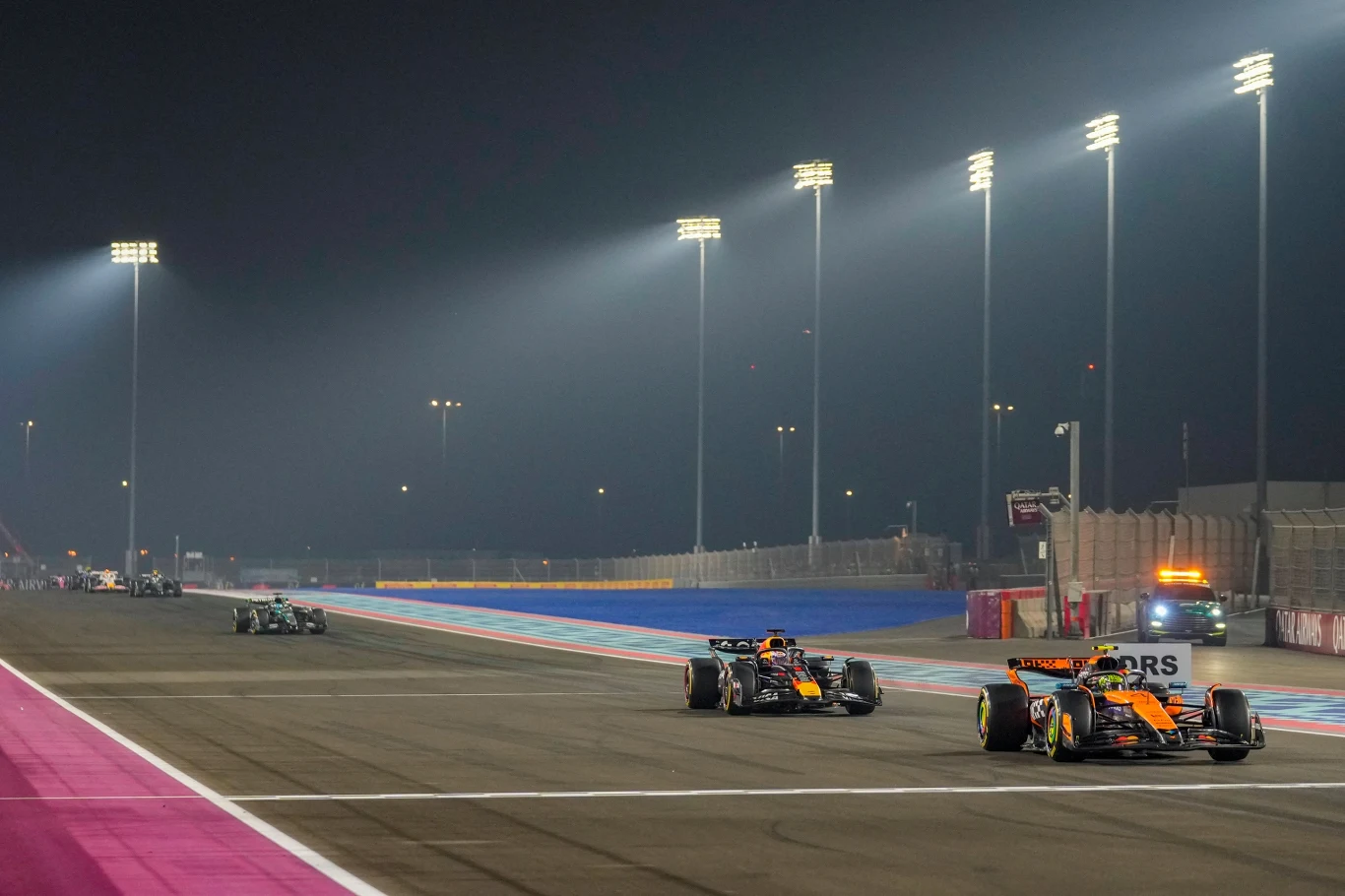 Oscar Piastri i Max Verstappen podczas Grand Prix Kataru Wyścigowe bolidy Formuły 1 rywalizujące na torze oświetlonym mocnymi reflektorami nocą, z kilkoma pojazdami na asfalcie, trybuny i elementy toru widoczne w tle.