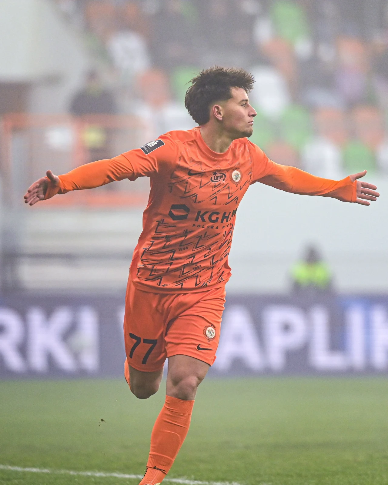Piłkarz w pomarańczowym stroju z numerem 77 świętuje zdobycie bramki, biegnąc z rozłożonymi ramionami po murawie stadionu podczas meczu piłkarskiego. W tle widoczni kibice oraz elementy stadionu.