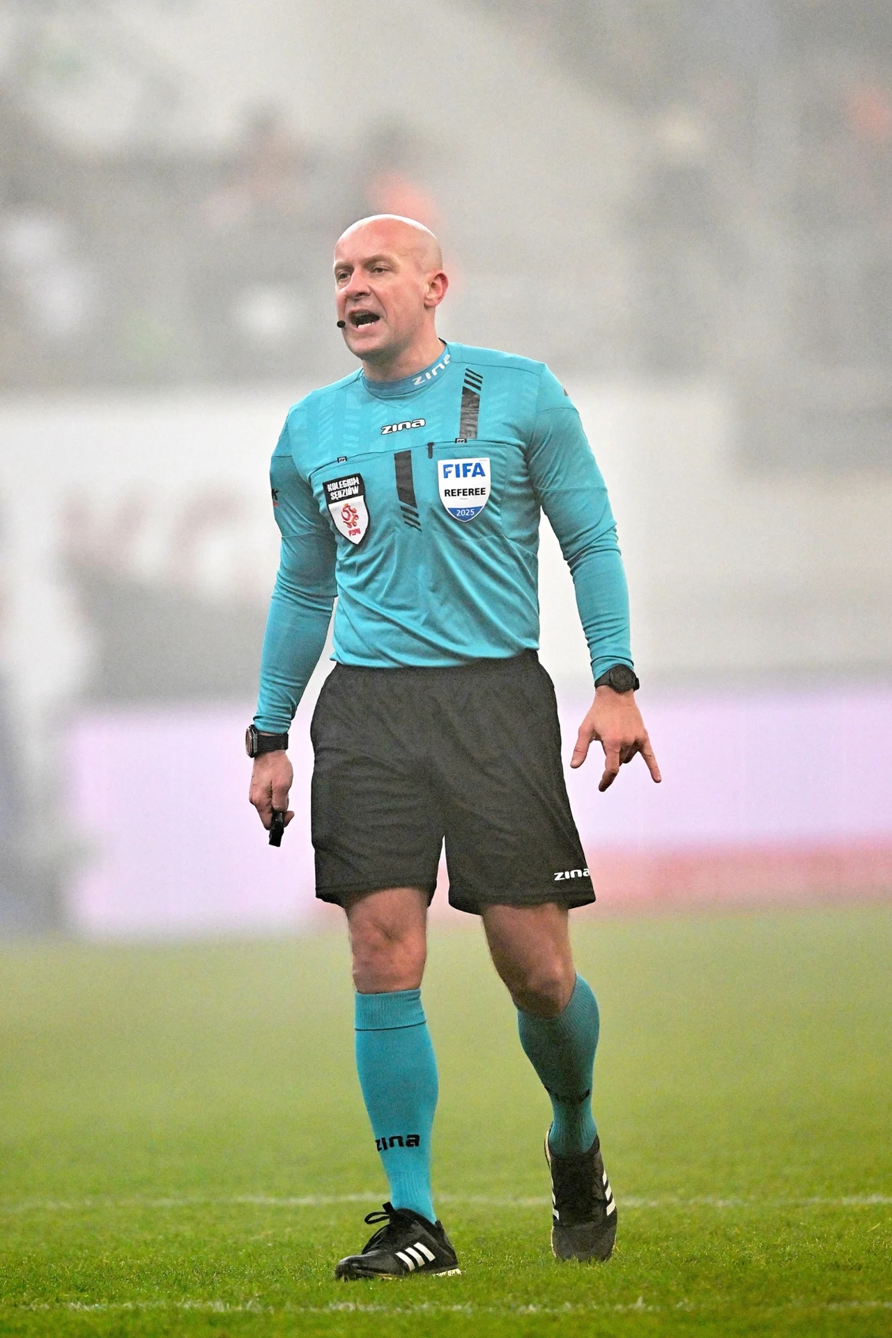 Sędzia piłkarski w niebieskim stroju, stojący na boisku podczas meczu, z gwizdkiem w ręce, w tle zamazany obraz stadionu i rozmyte postacie kibiców.