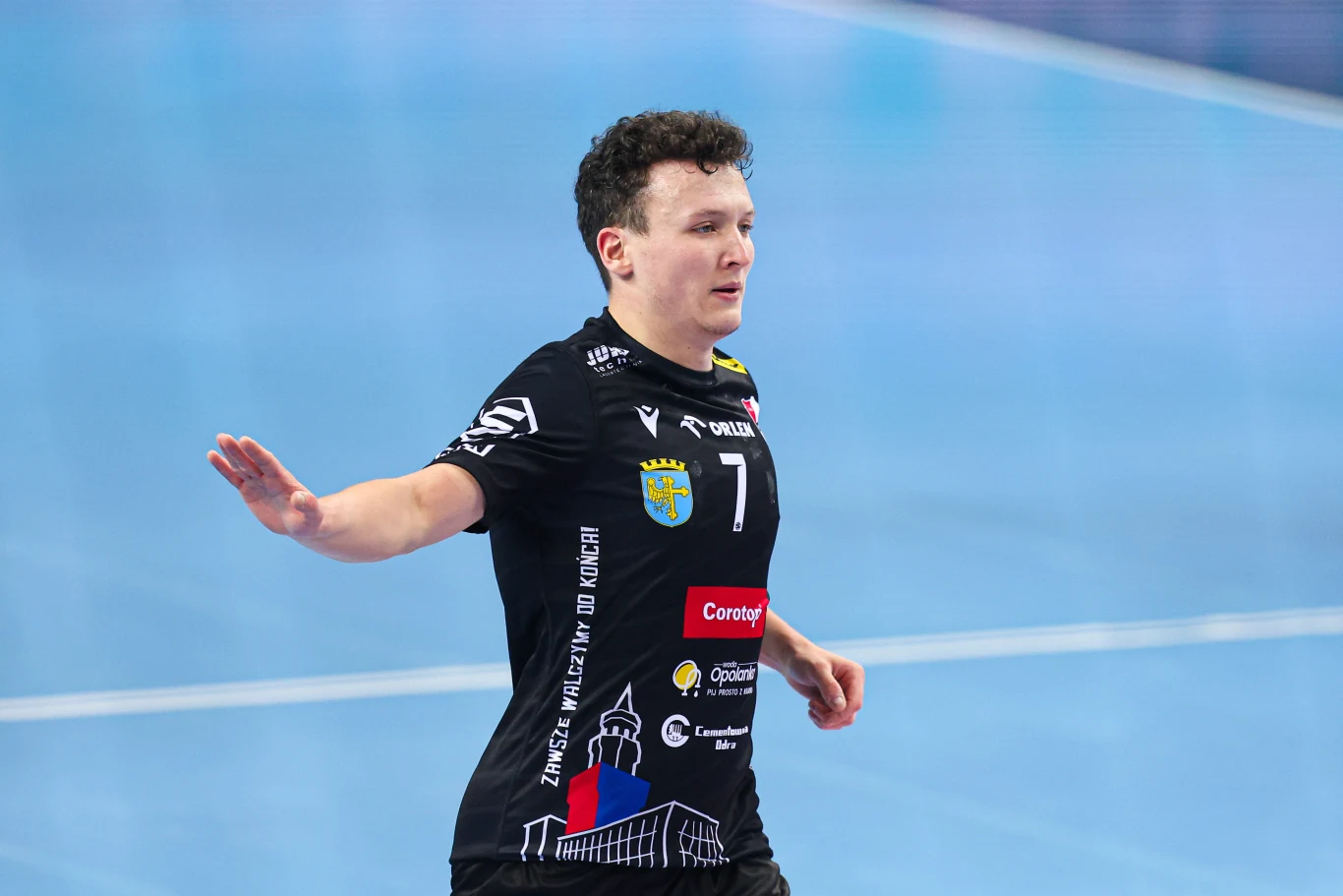 Mateusz Wojdan Zawodnik drużyny piłki ręcznej w czarnej koszulce z numerem 7 wykonuje gest ręką na tle niebieskiego boiska sportowego, widoczne logotypy sponsorów na stroju.