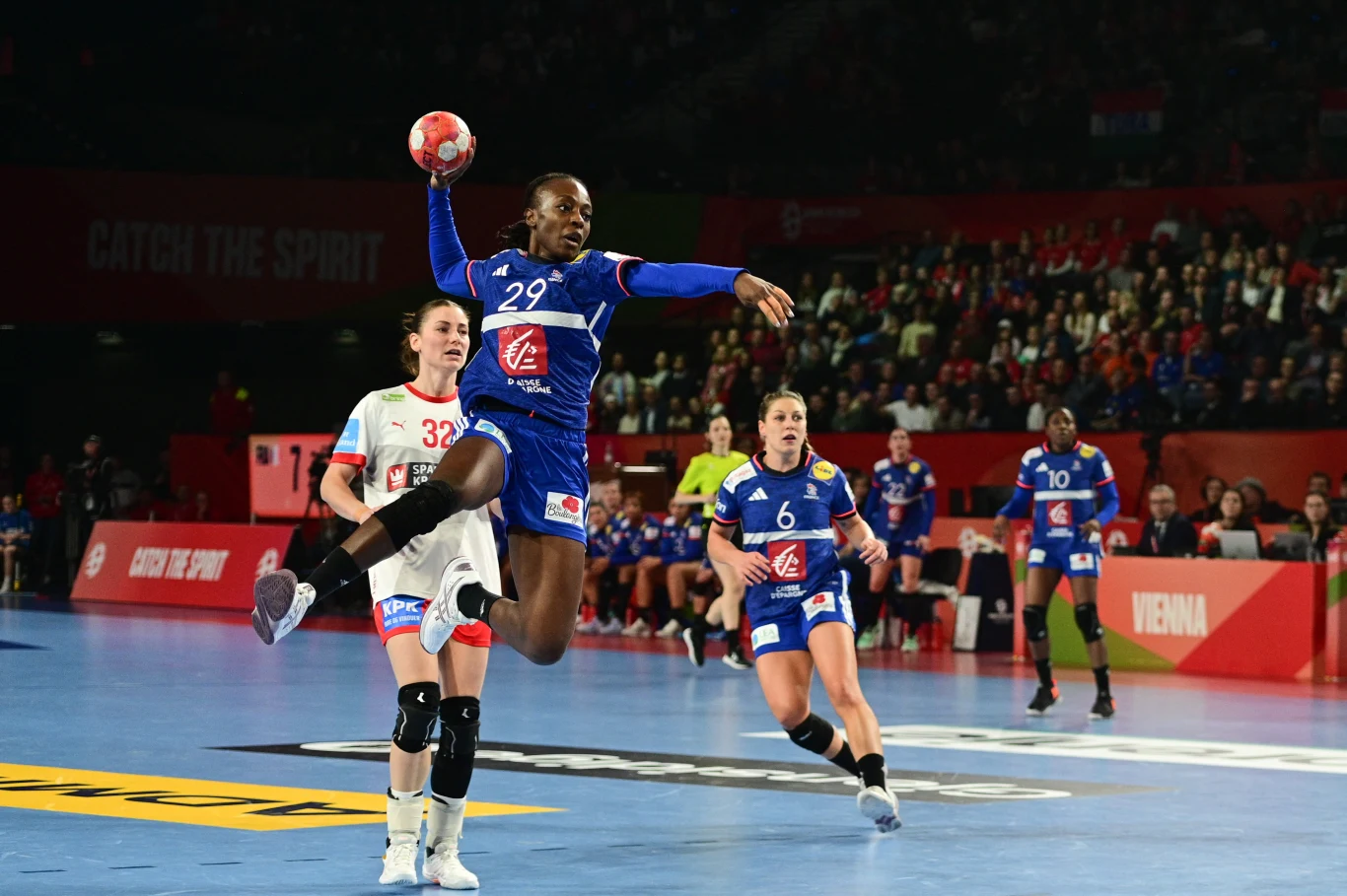 Zawodniczka w niebieskim stroju unosi się w powietrzu podczas rzutu do bramki w meczu piłki ręcznej, w tle trzy inne zawodniczki, kibice na trybunach i wyraźne światła hali sportowej.