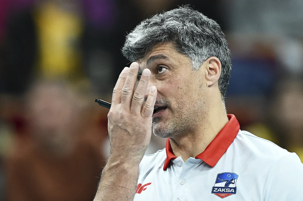 Andrea Giani, trener ZAKS-y Kędzierzyn-Koźle Mężczyzna z siwiejącymi włosami w białej koszulce z czerwonym kołnierzykiem w barwach klubu sportowego ZAKSA, podnosi rękę z długopisem w stronę twarzy, będąc w skupieniu.