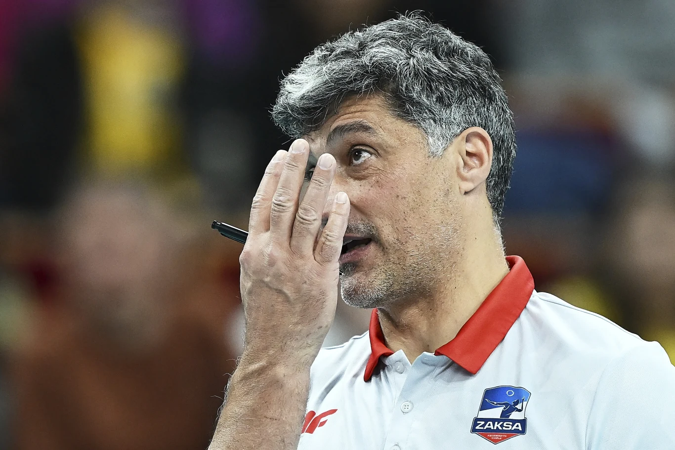 Andrea Giani, trener ZAKS-y Kędzierzyn-Koźle Mężczyzna z siwiejącymi włosami w białej koszulce z czerwonym kołnierzykiem w barwach klubu sportowego ZAKSA, podnosi rękę z długopisem w stronę twarzy, będąc w skupieniu.