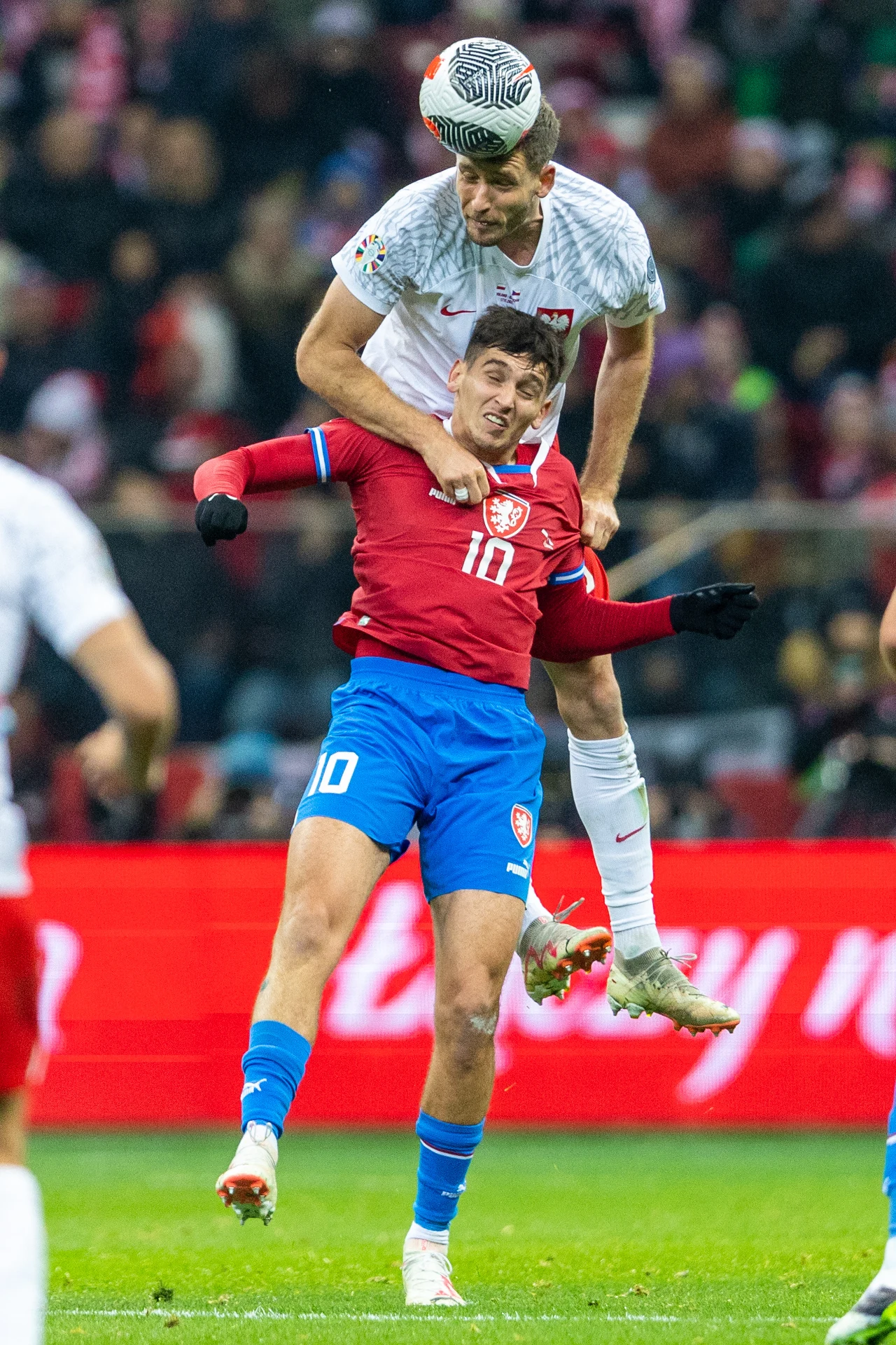 Dwóch piłkarzy walczy o piłkę w powietrzu podczas meczu, jeden z nich jest ubrany w czerwony strój i numer 10, drugi w biały strój z orłem na piersi, na tle trybun pełnych kibiców.