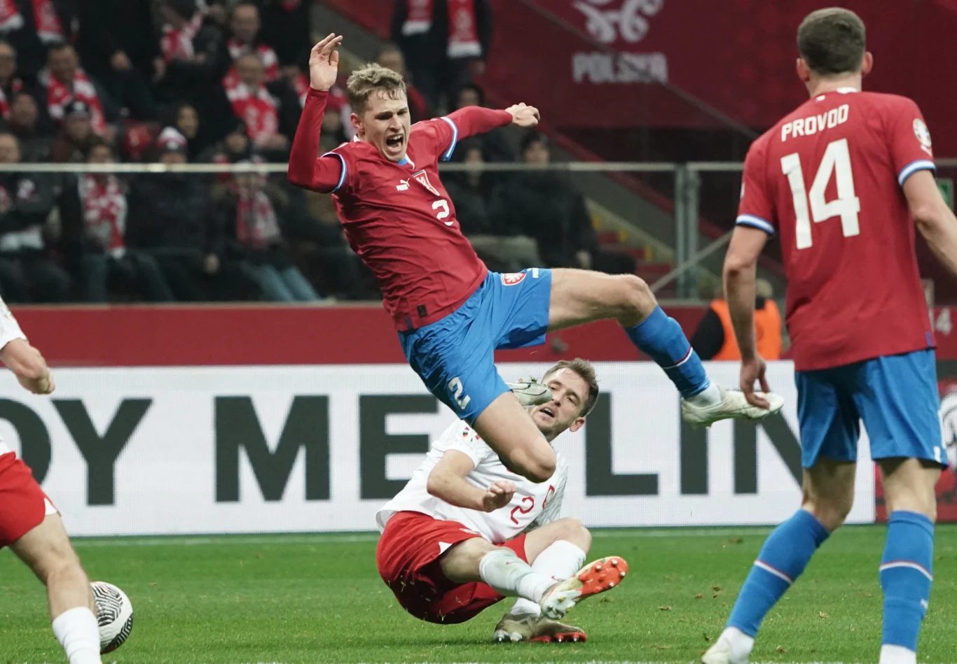 Dynamiczna akcja piłkarska podczas meczu, zawodnik w czerwonym stroju unosi się w powietrzu po faulu przeciwnika w białej koszulce, na trybunach widoczna publiczność kibicująca drużynom.