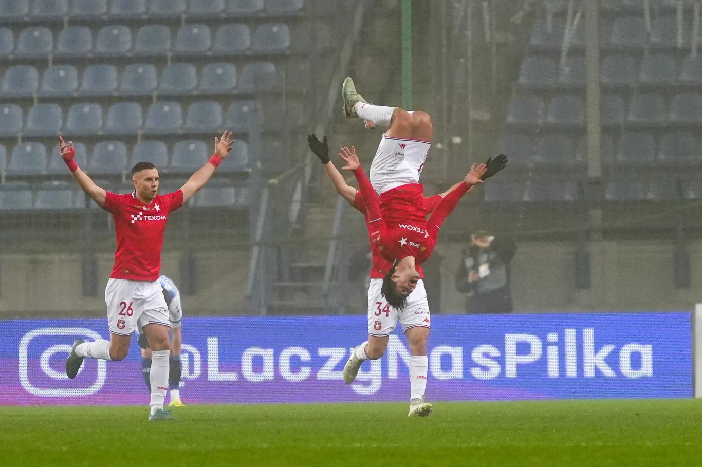 Tak Frederico Duarte świętował zdobycie bramki Zawodnik w czerwonej koszulce wykonuje efektowne salto w powietrzu celebrując zdobycie gola, obok niego drugi piłkarz cieszy się z podniesionymi rękami, w tle puste krzesełka stadionowe oraz reklama.
