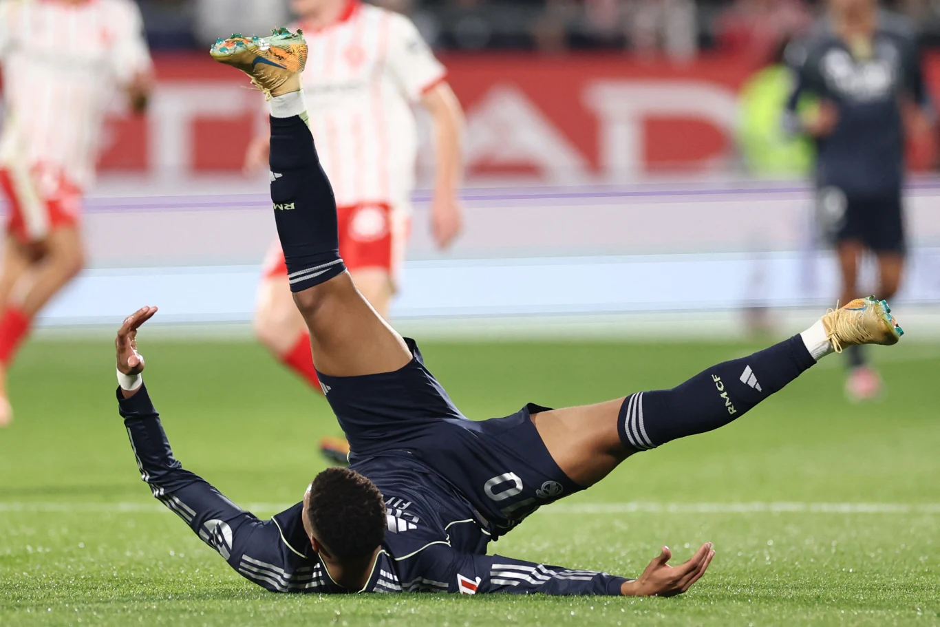 Piłkarz w ciemnym stroju leżący na murawie z nogami uniesionymi w powietrzu podczas upadku, w tle sylwetki zawodników w biało-czerwonych strojach oraz rozmyte elementy stadionu.