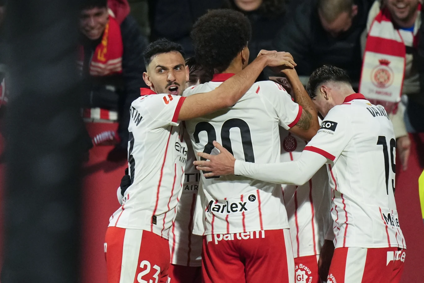 Grupa piłkarzy w białych koszulkach z czerwonymi elementami obejmuje się i świętuje zdobycie gola na stadionie, w tle widoczni kibice z szalikami.