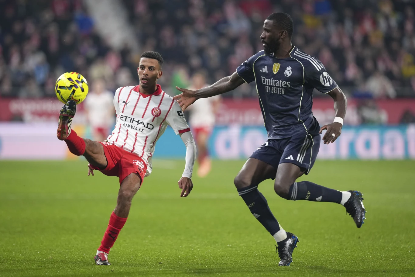 Dwóch piłkarzy walczy o piłkę podczas meczu piłkarskiego, jeden z nich kopie piłkę, drugi zbliża się próbując ją przejąć, obaj są w pełnym stroju sportowym na murawie stadionu.