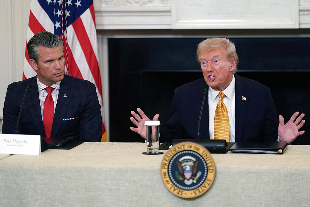 Sekretarz wojny Pete Hegseth i prezydent Donald Trump Dwóch mężczyzn siedzi przy stole konferencyjnym, jeden z nich zakłada garnitur i czerwoną krawat, drugi ma ciemny garnitur i żółty krawat. Na stole znajduje się mikrofon, szklanka wody oraz plakietka z pieczęcią Prezydenta Stanów Zjednoczonych. W tle w...