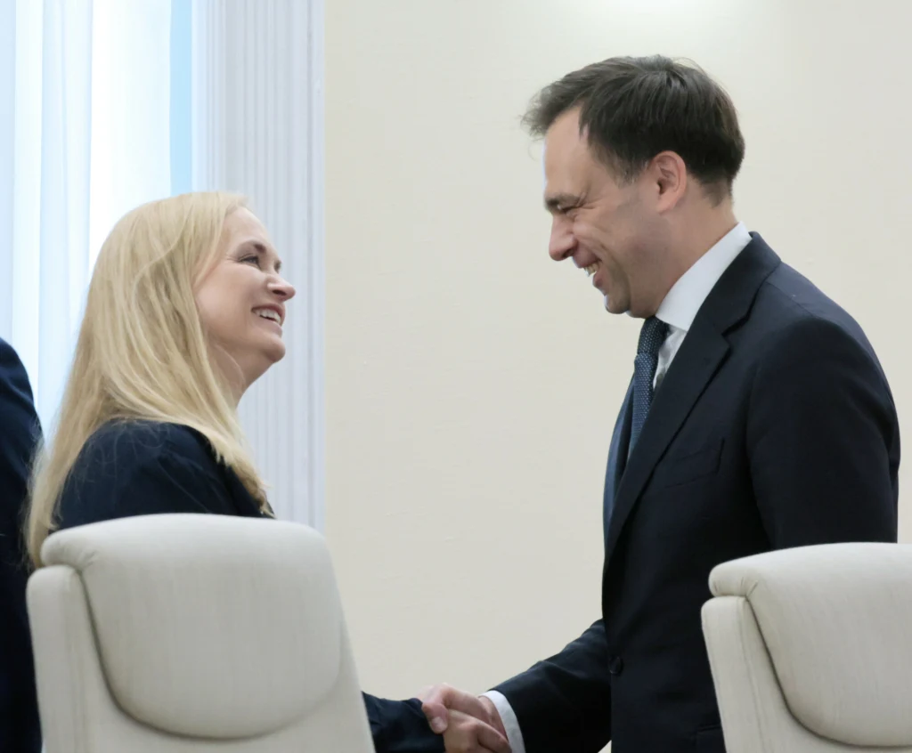 Uśmiechnięta kobieta i mężczyzna w garniturze podają sobie ręce, nawiązując kontakt wzrokowy, w jasnym i formalnym wnętrzu.