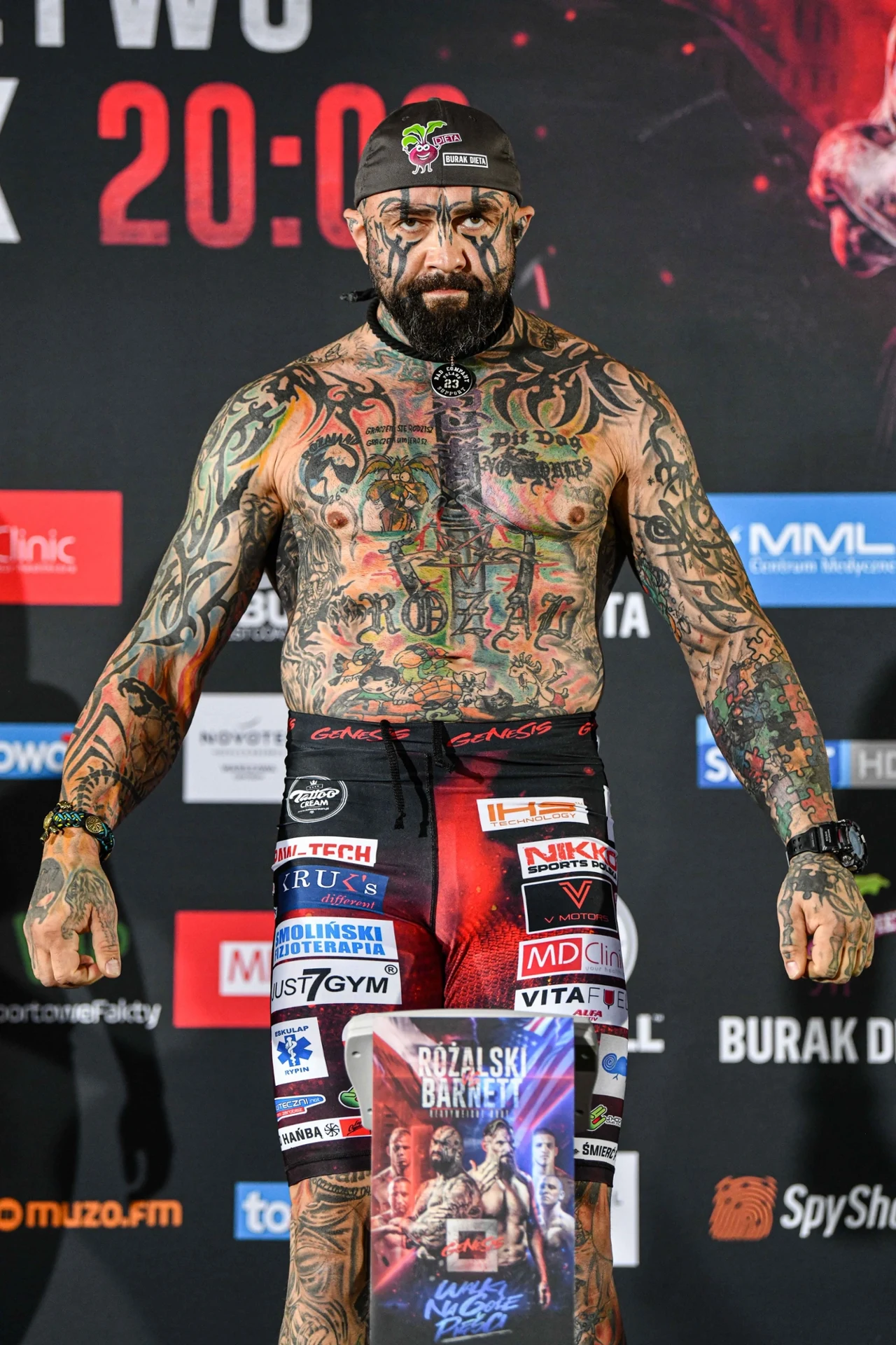 Mężczyzna o licznych tatuażach na ciele oraz twarzy stoi wyprostowany na tle planszy z logotypami sponsorów, ubrany w sportowe spodenki bokserskie pokryte naszywkami i reklamami.