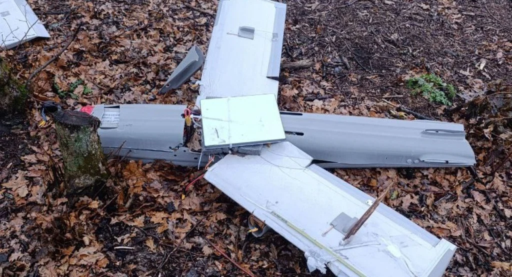 Rozbity dron leży wśród opadłych liści na leśnym podłożu, fragmenty kadłuba i skrzydeł są widocznie uszkodzone, wokół widoczne są drzewa i gałęzie.