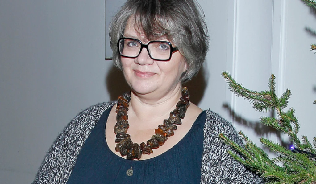 Dorota Zawadzka Kobieta w okularach i krótkich szaro-brązowych włosach, ubrana w ciemną bluzkę i sweter, z dużym naszyjnikiem z bursztynów, stoi obok gałęzi choinki.