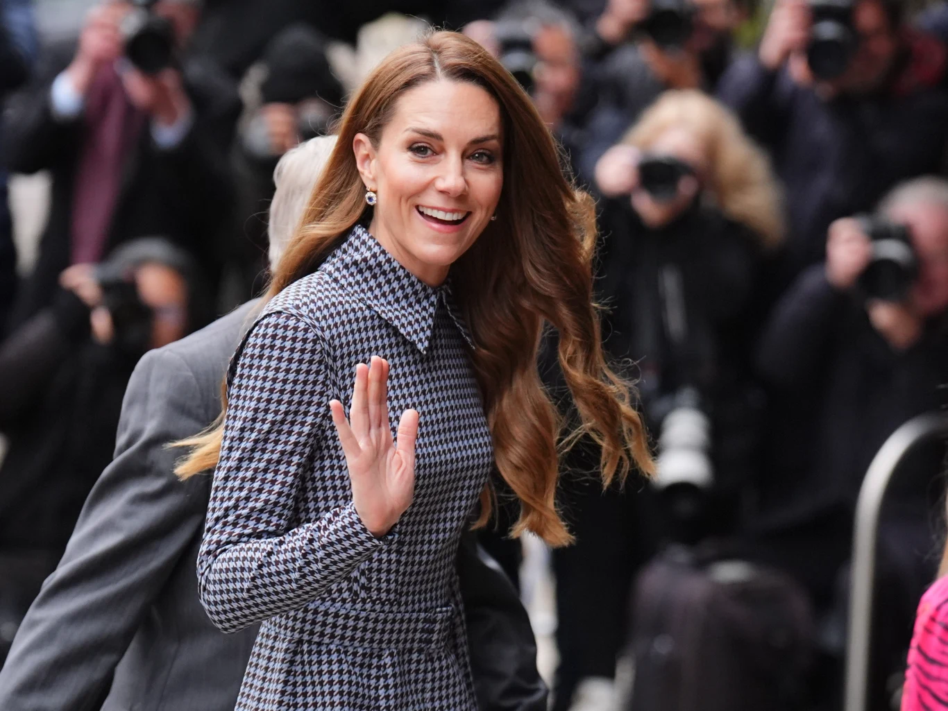 Księżna Kate zachwyciła stylizacją kolejny raz Uśmiechnięta kobieta z długimi, falowanymi włosami w eleganckim płaszczu w kratę macha do zgromadzonego tłumu fotoreporterów, w tle liczne osoby z aparatami fotograficznymi.