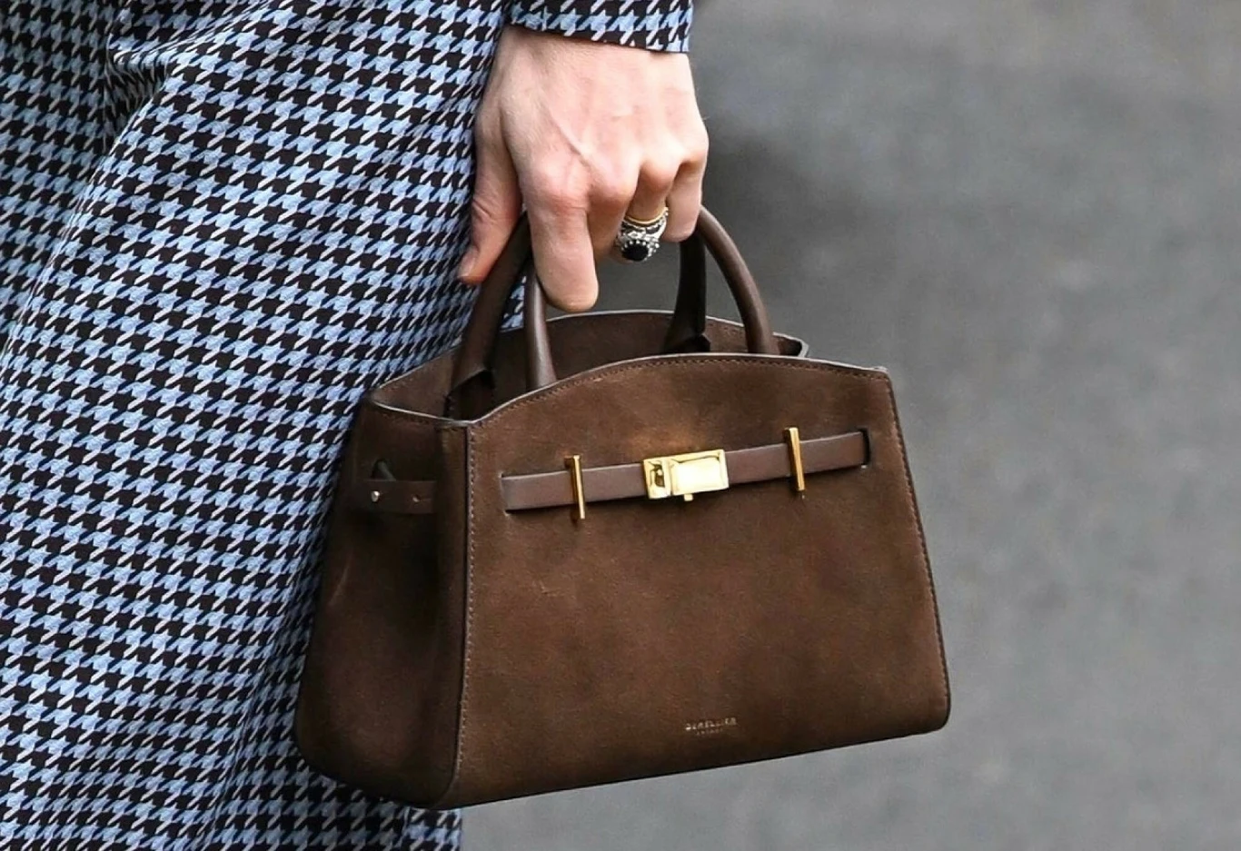 Torebka księżnej Kate przyciągnęła wzrok fanów mody Brązowa torebka z eleganckim zamkiem i złotymi elementami trzymana przez osobę ubraną w ubranie w niebiesko-czarną pepitkę.