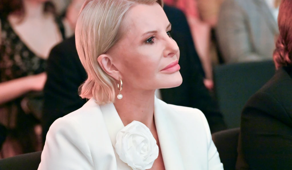 Joanna Racewicz Elegancka kobieta z krótkimi blond włosami w białym stroju ozdobionym dużym kwiatem, siedząca wśród publiczności, skoncentrowana na wydarzeniu.